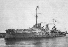 A large gray warship sits in harbor, wispy smoke billows lazily from two smoke stacks