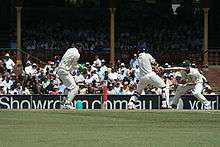 A cricket match being held. The batsman hits the ball and the other players try to catch it. The green field and the audience are visible in the far.