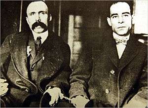 Black and white photograph of two men in suits and handcuffs, sitting down.