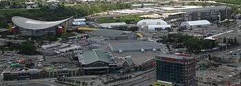 A downward-looking image of numerous buildings.  A large arena is situated to the left, behind a building with a green peaked roof.  To the right in the distance is a dirt race track.