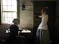 Living History interpreters portraying two young women of Salem. The seated woman is copying Bible verses using a quill.