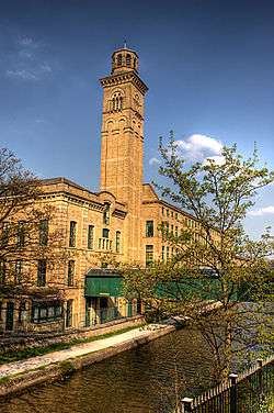 A three-story pale-red brick building along the left side of a small river. The building has a bell tower rising out of the middle of it.