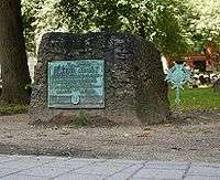 A rectangular, rough-hewn block of stone on the ground. There is a walkway in the foreground, grass and trees in the background. A weathered plaque on the stone reads: "Samuel Adams, Signer of the Declaration of Independence, Governor of this Commonwealth, A Leader of Men and an ardent Patriot."