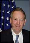 Smiling man with thinning hair wearing a suit and a blue tie with the US flag behind him