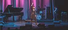 36-year-old Blasko is shown in centre of a stage. She holds a microphone in her right arm raised high above her head while singing into another microphone on its stand. She wears a dark dress and has a single-stranded necklace. On her extreme right is a piano with the player's head just visible as he leans towards the instrument. To her left and behind is a drummer at his kit. A guitarist is further to her left. Other stage equipment is visible. The fourth backing band member is out of shot.