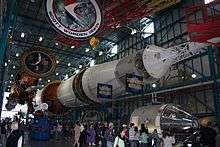 The Saturn V rocket is displayed horizontally, its engines at the left and the command module at its top on the right. Above it hangs large circular logos of each of the Apollo missions.