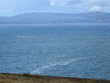 The attack site today, seen from a cliff above the bay. A small green wreck buoy is a few hundred metres away. A thin slick of oil is on the surface of the sea.