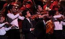 five children dressed in black and white play violins standing in front of seated adults in orange jackets