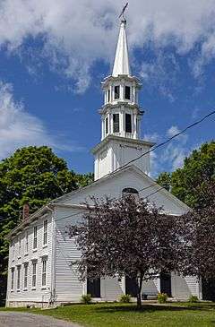 A white church with a tall spire, seen at a slight three-quarter angle from the front under a blue sky with clouds. There are some trees behind it and shorter ones in front.