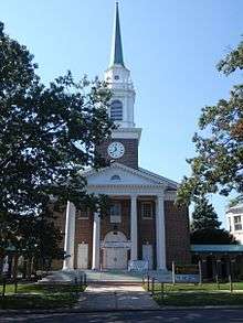 The front façade of a nineteenth-century brick church building with columns and Greek Revival pediment, cupola, and steeple