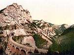 A photochorm picture of the a curving railroad bridge coming out of a tunnel bored into a large mountain.  In the background another bridge and mountains are visible.