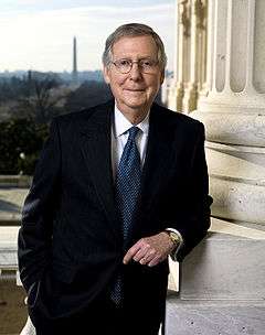 A man in his late sixties with graying hair and glasses leaning against a column and wearing black suit, white shirt, and blue tie