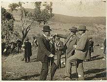 Three middle-aged men with short beards in formal suits and hats standing in open hilly field with single tree nearby