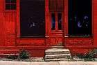  oil painting of a 200 year old building with 2 large pane-glass windows and 2 doors, steps, weeds—painted deep red with black windows mirroring blue sky