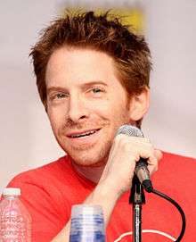 A man with red hair, smiling slightly and sitting behind a microphone