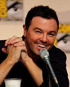 A Caucasian male with black hair and stubble, along with a black shirt. His hand is placed under his chin and a microphone is in front of him. A vague simple is marked behind him.