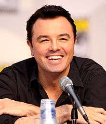 A man with black hair and a black shirt, leaning forward, smiling into a microphone