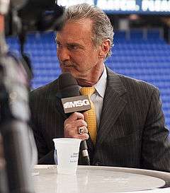 A grey-haired man in a black suit and yellow tie looks to his right, a microphone marked "MSG" held to his mouth.