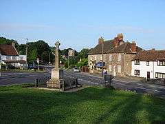 Stone cross surrounded by railings on grass area in front of roads and houses.