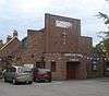 A brick building with an angular façade consisting of stepped brick walls above an entrance porch with a wooden door in a recess.  A crucifix and the words "SHOREHAM FREE CHURCH" are on the façade.