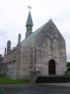 A grey-white stone chapel seen from the northwest, with a central spirelet with a cross. There is an elaborate carving of the Crucifixion over the west door, a tall pinnacle at the corner, and elaborate stone tracery in the windows along the side