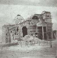 Photograph showing a domed masonry building with its left corner and most of its facade reduced to rubble and only one tower on the right and half of the pedimented porch intact