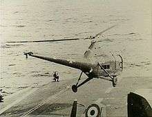 The forward section of an aircraft carrier's flight deck. A helicopter with United States markings and the letters "UP-28" painted on the side is hovering just above.