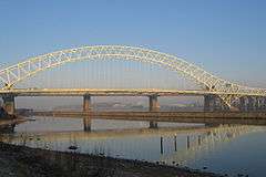 In the foreground is the Manchester Ship Canal with a portion of shore and its wall beyond; behind this is a grey-coloured arch carrying a roadway with the piers of the railway bridge beneath; under the arch are factories on the far side of the river