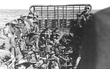 About forty troops wearing slouch hats and carrying rifles packed into a landing craft travelling across open water, viewed from the stern towards the bow ramp.