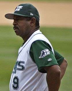 A man wearing a white baseball uniform with dark green sleeves bearing a white "M" and a dark green cap with a white "M" on the front stands on a baseball field