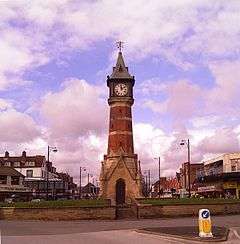 Octagonal brick and limestone tower on a square limestone base, designed like a Gothic church porch. The tower is capped with a square lantern, a white clock face on each side, heavy with Roman numerals. The towering roof is capped with a gold-coloured weathervane. The viewpoint is on the short coast road called Tower Esplanade; behind the tower is the row of tourist-trap shops in Lumley Road. The tower dominates the picture, standing out of a partly blue sky mostly filled with white, purplish, clouds. There is a ring of brick flowerbeds ringing the roundabout where the tower is. The photo was taken in May, the flower beds are just green.