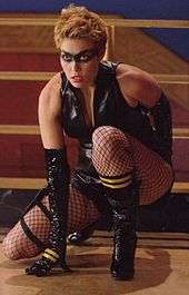 A blonde woman sits crouched on one knee. She is wearing a black painted-on mask, with a black leather vest. She is also wearing black, elbow-length gloves, fishnet stockings, and knee-high black leather boots. The boots and gloves both have two gold, horizontal stripes. In the background is the railing to a staircase.