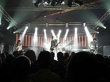 The Smashing Pumpkins perform on a back-lit stage dressed in white. From left to right: Ginger Pooley—a brunette Hispanic woman in her 20s wearing a sequined dress—looks at her bass guitar, Billy Corgan—a bald, middle-aged Caucasian man with a white jumpsuit and a black-and-white striped T-shirt—plays guitar, Jimmy Chamberlin plays a silver drum kit obscured behind Corgan, and Jeff Schroeder—a Korean male in his 30s with mid-length brunette hair—looks at his guitar while playing.