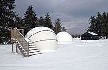 Two white observatory domes in a snow-covered field, wooden stairs are beside one dome and a cabin and trees are in the background