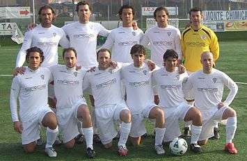 Six men are down on one knee on a grass football field. Five men are standing behind them. The man standing on the top right is wearing a yellow top, with the letters "BROS" across his top. The other ten are wearing similar tops, but in white.