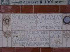 A tablet formed of five tiles of varying sizes, bordered by yellow and blue flowers in an art nouveau style. The tablet reads "Solomon Galaman, Aged 11 died of injuries, Sept 6 1901 after saving his little brother from being run over in Commercial Street, 'Mother I saved him but I could not save myself.'"