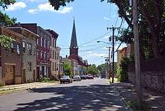 A view down a city street with a group of rowhouses of differing heights and colors on the left. A tall church spire is visible in the distance. Along the front lawn, which rises slightly in front of the camera, is a line of six s