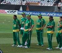 Six females in green cricketing outfits standing on the outfield looking at the presentation stage