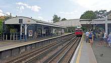 Two sets of railroad tracks, both with powered third rails and middle guide rails, between elevated concrete platforms with white curved wooden canopies. In the background is a bridge with curved solid white wooden walls. A sign on the far platform, at left, says "Kew Gardens". A red train is pulling into the station's near platform.