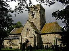 A stone church seen from the south, with a central tower in front of which is a two-storey gabled porch.  To the left is a short nave, and to the right an apsidal chancel