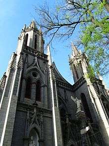 Two ornate spires of grey stone, with a tree branch partially in leaf at right, seen against a blue sky at a sharp upward angle