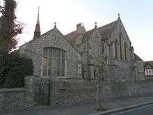 A low-roofed, wide, cobbled flint church with several sections, facing a road behind a wall of the same material. On the right, partly obscured, is an apse with plain stone-dressed lancets; next to it is the body of the church, with tall round-headed windows.  In the foreground is a spirelet and a low extension with a five-light window.