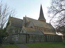 Side view of a long flint-built church with several parts, each with a different roofline.  A spire is visible behind the body of the church.  All windows are framed by brickwork.