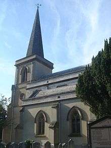 The tower, spire and part of the body of a dull grey church building. The stepped tower has one round window and one pointed-arched louvre, and is topped with a spire and weather-vane.  Two recessed two-light lancet windows with quatrefoils are on the near side, which is partly obscured by a tree.