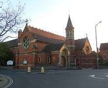 Three-quarter view of an ornate brick building with pale stone dressings and recessed lancet and round windows. A rose window dominates the left-hand side.  Perpendicular to its right, there is an arched entrance porch; beyond that is an octagonal tower with louvre-style openings below a conical spire.