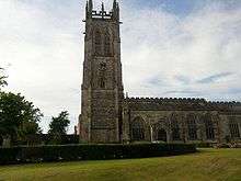 A Gothic church with a tall west tower surmounted by a complex parapet and pinnacles