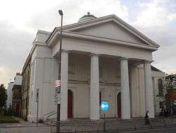 A Classical style, stuccoed building whose façade is dominated by four tapering columns supporting a pediment.  The side wall is yellow brick.  Partly hidden behind the columns are two red round-headed doors.  Above the pediment is a partly hidden cupola.  A modern extension is partly visible to the right.