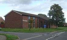 Distant three-quarter view of a modern L-shaped brick building with three ground-to-roof windows on the longer side and a blank wall on the shorter side.  The tiled roof extends to just above ground level on the right side to cover an extension with two tall windows.  A tall tree in full leaf stands next to this.