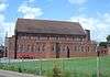 Side view of a tall, plain red brick church with an entrance porch at the left end and several uneven projections from the near side.  The porch has a stone statue and an arched entrance door.  The tiled roof has stone ends, both of which have small stone crosses.  The lower level of the longer side has 15 unevenly spaced but identical round-headed windows; above this and immediately below the roofline are another eight windows of the same type, which are evenly spaced.