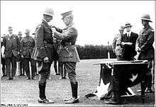 a black and white photograph of a male in uniform with peaked cap pinning a medal on the jacket of a second male in uniform wearing a pith helmet